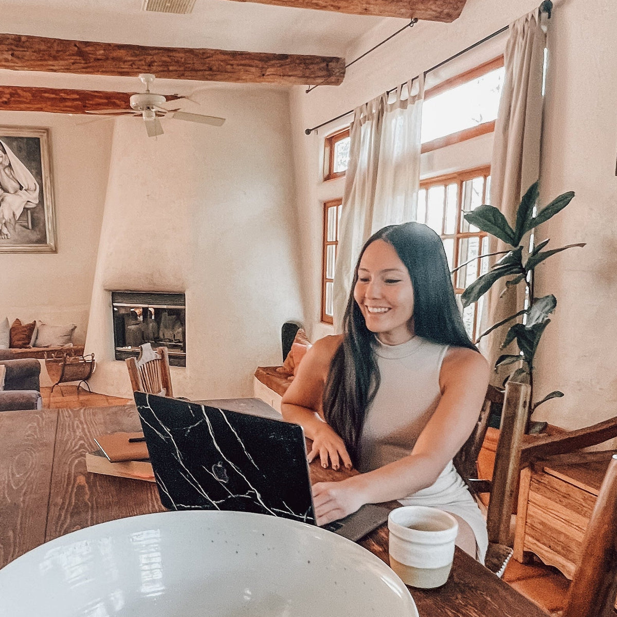 A woman sits at a wooden dining table in a rustic home interior, using a laptop. A large white bowl is prominently featured in the foreground on the table, with wooden chairs, exposed wood beams, and framed wall art visible in the room.