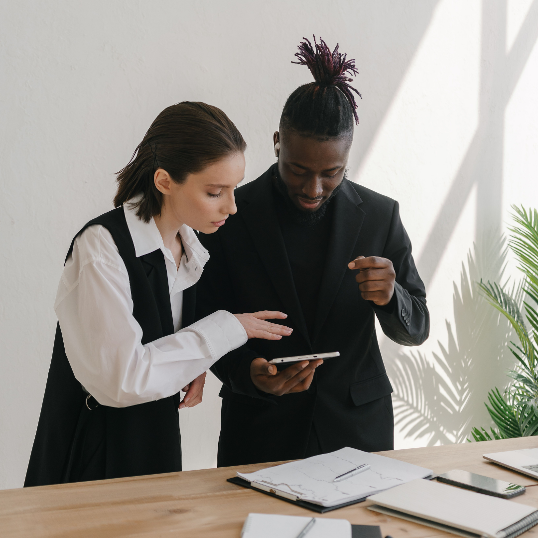 A man and a woman in business attire stand together at a wooden desk, looking at a tablet screen in a bright office with natural light and a plant in the background. doing their content directive for their small business.